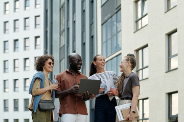 Poster - Young cheerful intercultural employees chatting and laughing outdoors while enjoying coffee break on their way to office along buildings