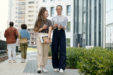 Poster - Two young women in casualwear discussing financial papers while walking to office along modern buildings against intercultural couple