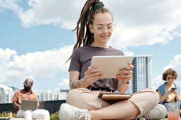 Poster - Happy young female student or employee looking at tablet screen while watching online video or working over new project in open air