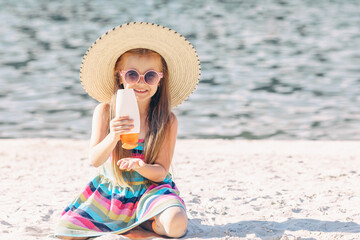 Cute happy little blonde hair girl in straw hat using sunscreen cream on a sunny sandy beach. Summer vacation concept