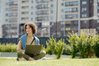 © pressmaster - Young smiling woman with laptop sitting on green grass in park on sunny summer day and looking aside while enjoying rest and watching video