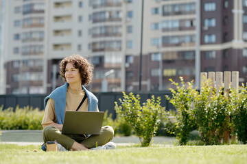 Poster - Young smiling woman with laptop sitting on green grass in park on sunny summer day and looking aside while enjoying rest and watching video