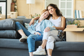 Wall Mural - Two happy young girlfriends in casualwear sitting on comfortable couch in living room and enjoying rest after relocation to new apartment