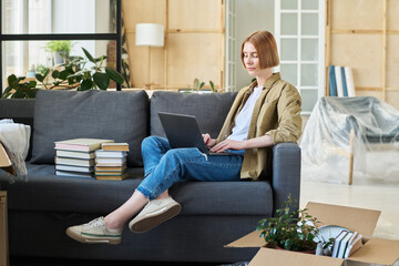 Wall Mural - Young serious businesswoman or student typing on laptop keyboard while sitting on couch with stacks of books and networking in living room