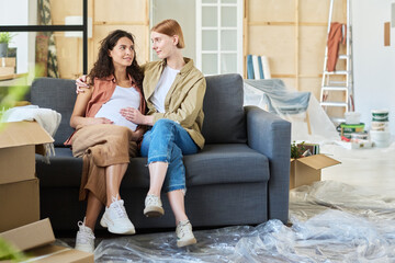 Wall Mural - Young woman sitting on couch next to her pregnant girlfriend, looking at her and touching her belly while relaxing in living room after relocation