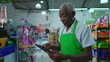 © Marco - African American senior employee of supermarket using tablet device standing inside grocery store, job occupation of a black older person at workplace, checking inventory