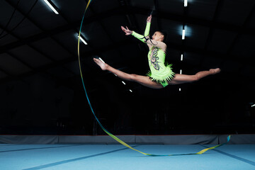 Wall Mural - Jump, rhythmic gymnastics and woman in gym with ribbon, creative sport or action, performance or fitness. Competition, athlete and female gymnast, dance and art with body, routine and energy at arena