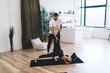 © BullRun - Woman doing yoga position with help of trainer in living room