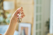 Hand of young woman holding metallic key with house shaped trinket hanging on ring in front of camera against interior of living room