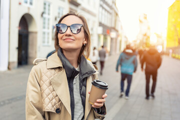 Portrait young beautiful happy smiling woman drinking coffee to go tea in eco sustainable paper cup wear biege trench coat enjoy walking european city street. Stylish female person urban lifestyle