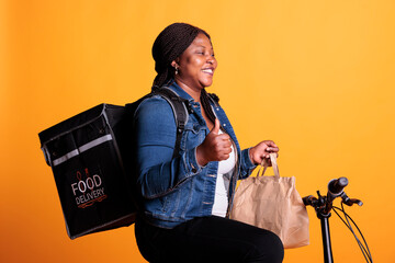 Wall Mural - Positive courier holding paper bag full with takeaway food order delivering to customer during lunch time, worker doing thumbs up gesture at camera. Deliverywoman using bike as transportation