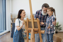 Happy Ladies And Senior Man Laughing At Image On Canvas While Standing Behind Easel In Home Art Space. Smiling Mother Taking Break From Serious Painting When Having Fun In Studio With Dad And Kid.