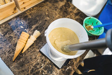 Closeup Of Professional Blender Whisking Ice Cream. Bucket Of Bright Yellow Ice Cream In Bucket On Brown Kitchen Counter With Ice Cream Cones And Plastic Spatula. Ice Cream Production Process Concept