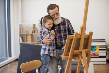 Caring grandparent cuddling little child with paintbrush while doing easel-type portrait in home art studio. Grey-haired mature man sharing creative skills with active granddaughter on weekend.