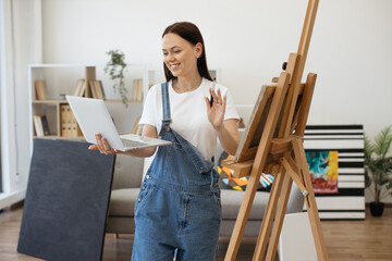 Wall Mural - Cheerful woman having group video call on wireless computer and waving hand to greet friends in bright spacious room. Pretty brunette participating in online school for young artists.