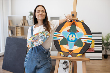 Wall Mural - Portrait of confident female artist holding palette with paintbrush in hands and smiling at camera. Casually dressed dark-haired woman posing near easel in creative art studio.