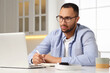 © New Africa - Young man with laptop writing in notebook at desk in kitchen. Home office