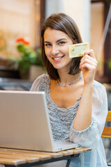 Woman using credit bank card laptop computer while transferring money, purchases online shopping order food delivery booking hotel room outdoors. Girl tourist sits at table in city cafeteria. Vertical