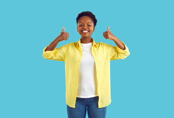 Joyful young ethnic woman giving two thumbs up recommending or approving great choice. African American woman with beautiful smile in casual clothes demonstrates okay gesture on light blue background.