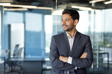 Young successful arab businessman with crossed arms smiling and looking out the window, financier inside office at workplace standing satisfied with achievements.