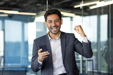 Portrait of successful financier investor, hispanic man smiling and looking at camera, received online win message, man holding hand up gesture of triumph and winner, inside office at workplace.