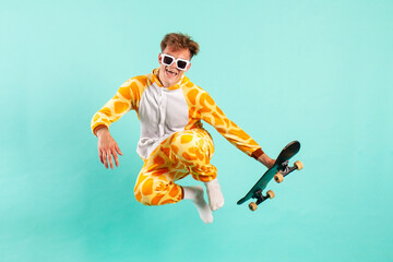 Wall Mural - young funny guy in orange giraffe pajamas jumps and flies with skateboard on blue isolated background