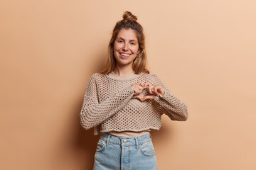 Woman extends her hands forming heart shape as symbol of love and connection her genuine expression conveys warmth and compassion creating heartfelt moment dressed casually poses against brown wall