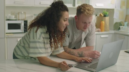 Medium shot of young family couple leaning on kitchen table, browsing marketplace website on laptop and credit card while doing online shopping together at home