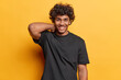 Positive emotions. Studio shot of young happy smiling broadly Hindu man standing isolated in centre on yellow background wearing casual black tshirt keeping left hand raised on neck right down