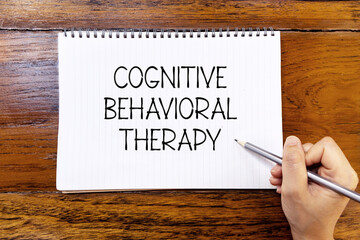 Cognitive behavioral therapy handwriting text on blank notebook paper on wooden table with hand holding pencil. Business concept and legal concept about Cognitive behavioral therapy.