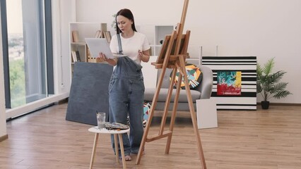 Wall Mural - Female professional painter having video conference using portable computer in comfy flat with amazing panoramic view. Attractive female in casual outfit having online art class with beginners.