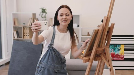 Wall Mural - Positive lady in every day clothes making selfie on modern smartphone and waving hand near wooden easel in creative art studio. Cheerful female spending weekend by doing favourite hobby.