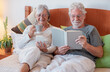 © luciano - Smiling senior couple resting in bed, man reading a book, woman using phone drinking a coffee cup.