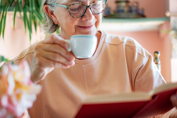 Poster - Smiling senior woman sitting at cafe table reading a book or studying enjoying an espresso coffee. Elderly lady holding a white coffee cup