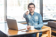 © khosrork - Portrait of strict bossy young bearded handsome man freelancer in blue jeans shirt working on laptop, pointing at camera, selecting you. Indoor shot near big window, cafe background.