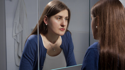 Sad young brunette woman looking at herself in mirror. Concept of depression, stress, mental illness and problems, loneliness and frustration.