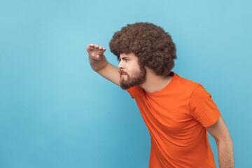 Side view of man with Afro hairstyle wearing orange T-shirt looking far away at distance with hand over head, attentively searching for bright future. Indoor studio shot isolated on blue background.