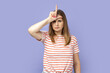 Portrait of blond woman wearing striped T-shirt showing looser gesture holding fingers near forehead, sad because of silly mistake. Indoor studio shot isolated on purple background.