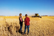 © Designpics - A view from behind of a mature farm woman standing in a field working together with a young woman at harvest time, while a combine is working in the background; Alcomdale, Alberta, Canada