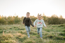 Daughter, Son Hold Hands And Run In Green Grass In Field At Sunset. Happy Children Walking Spending Time Together In Nature. Concept Family Holiday Outdoors. Kids Playing In Mountains On Autumn Day.