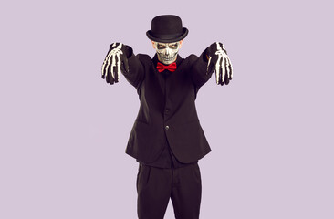 Skeleton man in Halloween costume looks ominously at you while standing in zombie pose. Young man with painted skull on his face stands in black suit with outstretched arms on lilac background.