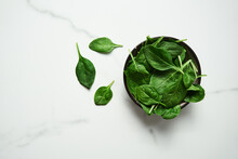 A Bunch Of Fresh Baby Spinach Leaves In The In A Black Ceramic Bowl On White Marble Background. Top View And Flat Lay Photo With Copy Space