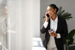© Prostock-studio - Happy black woman in suit having phone conversation and drinking coffee, female boss standing in office near window