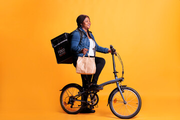 Wall Mural - African american delivery woman holding paper bag while delivering lunch order to client using bike as transportation. Restaurant courier carrying takeout thermal backpack, takeaway service concept