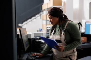 Wall Mural - Stockroom supervisor looking at inventory report on computer, working at merchandise quality control in warehouse. African american employee preparing customers orders for shipping in storehouse