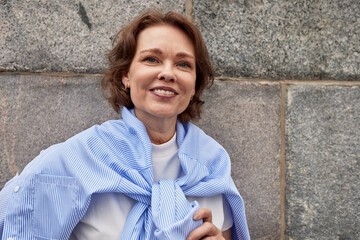 Wall Mural - Close up of attractive senior woman against the stone wall background. A middle-aged brunette in white t-shirt with blue shirt draped over the shoulders smiles happily. Active lifestyle for elderly.