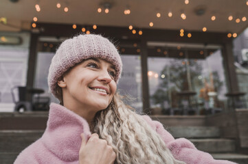 Fashionable mature woman in a pink knitted beanie hat and stylish coat outdoors in a cafe in autumn. Middle aged woman positive and smiling with pigtails in the city. Blur and selective focus