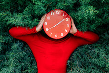Man Hiding Face With Red Clock Against Green Bushes