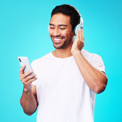 Poster - Headphones, music and Asian man with smartphone for listening in studio isolated on a blue background. Radio, smile and person with mobile, sound or hearing podcast, audio or social media on internet