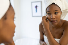 African American Woman Wearing Towel On Head Cleansing Face And Looking In Mirror In Bathroom